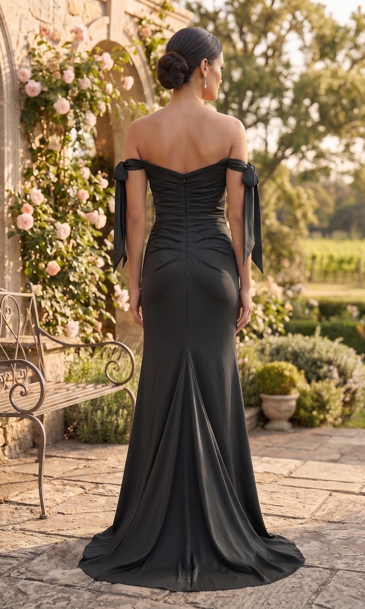 A woman stands outside on a stone patio, facing away, wearing the Chicas DM8010 Long Prom Dress—an off-the-shoulder, floor-length black gown with a thigh-high slit—set against a backdrop of a garden and blooming rose bushes.
